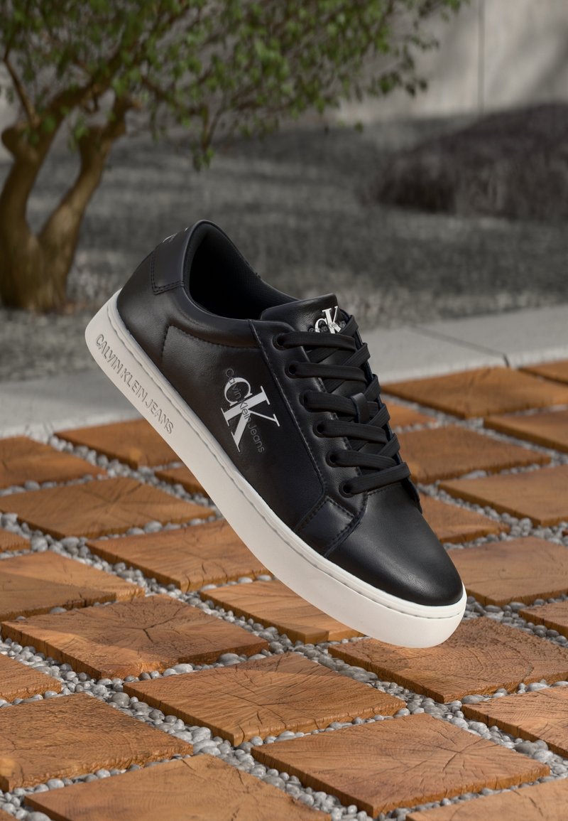 Svart lädersneaker med vit gummisula, med snörning och Calvin Klein-logga. Positionerad ovanför en yta av trä och grus.