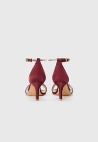 Burgundy satin korkeakorkoiset kengät nilkkahihnalla, avokärkiset, ja koristeelliset metalliset yksityiskohdat sivuilla. Kapea stiletto-korko.