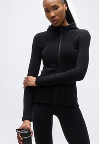 Γυναίκα με μαύρο αθλητικό μπουφάν με φερμουάρ και κολάν, κρατώντας ένα μαύρο μπουκάλι νερού με λαβή, σε απλό φόντο.