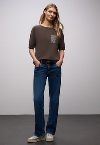 Braunes kurzärmeliges Hemd mit einer gemusterten Tasche, kombiniert mit blauer Jeans und hellen Sneakers, stehend vor einem neutralen Hintergrund.