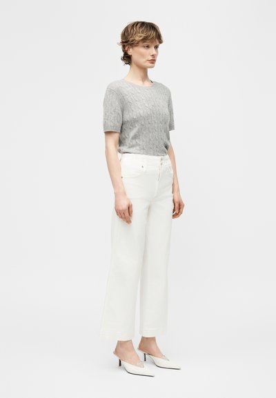 Γυναίκα που στέκεται φορώντας γκρι πουλόβερ με κοντά μανίκια και πλεκτό σχέδιο, λευκό παντελόνι με φαρδιά μπατζάκια και λευκά μυτερά παπούτσια με χαμηλό τακούνι kitten heels.