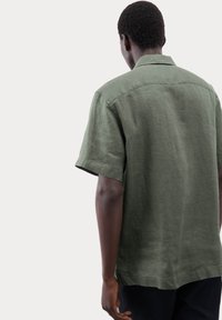 Korte mouwen, olijfgroene shirt van linnen. Heeft een kraagje, rechte zoom en een ontspannen pasvorm met zichtbare stikdetails.