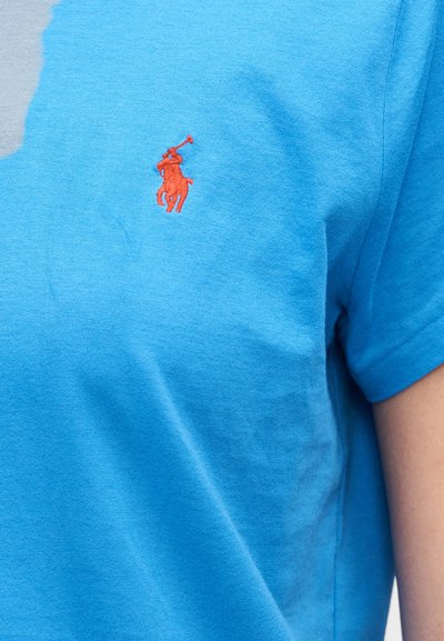 Šviesiai mėlynas medvilninis marškinėlis trumpomis rankovėmis. Kairėje krūtinės pusėje yra mažas raudonas siuvinėtas logotipas su arklio ir raitelio atvaizdu.