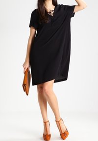 Robe noire à manches courtes avec une coupe ample et un décolleté lacé. Accompagnée d'une pochette marron et de chaussures à talons blocs orange.