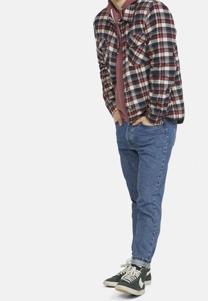 Personne portant une chemise à carreaux rouge et bleu marine par-dessus un sweat à capuche bordeaux, un jean bleu et des baskets vert foncé avec des lacets blancs, debout les mains dans les poches.