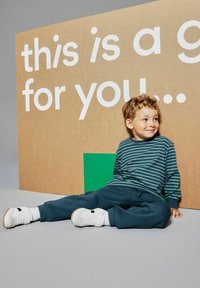 Młody chłopiec w pasiastym podkoszulku i ciemnych spodniach siedzi na podłodze, uśmiechając się, a w tle widnieje duży kartonowy znak z napisem "to jest prezent dla Ciebie".