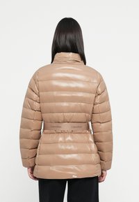 Pufová bunda v lesklom svetlohnedom materiáli. Má horizontálne prešívanie, vysoký golier a opasok s logom Calvin Klein v oblasti pásu.