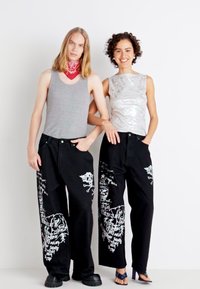 Jeans noirs avec des imprimés graphiques blancs, débardeur gris et top sans manches argenté ; le modèle à gauche porte un bandana rouge, les deux portent des chaussures contrastantes.
