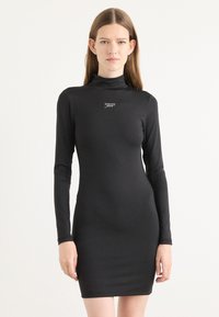 Svart langermet kjole laget av stretchy materiale. Har høy halslinning og en liten logo på brystet. Tett passform med minilengde.