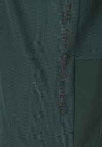 Donkergroene stof met een gestructureerd ruitpatroon. Bevat de bedrukte tekst "DE OFF-ROAD HELD" in het zwart langs de zijkant. Glad oppervlak, casual ontwerp.