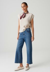 Cremfarbener, kurzärmeliger Oberteil kombiniert mit hochtaillierten, weit geschnittenen blauen Jeans. Leichter, glatter Stoff. Das Model trägt einen burgunder- und weißgestreiften Schal sowie cremefarbene Ballerinas.