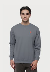 Giovane uomo con capelli scuri che indossa un semplice felpa grigia con un piccolo logo rosso sul lato sinistro del petto, in piedi davanti a uno sfondo bianco uniforme.