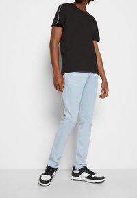 Camiseta de algodón negra con mangas cortas, jeans ajustados de color azul claro y zapatillas negras y blancas. Detalle del logo destacado en la manga.