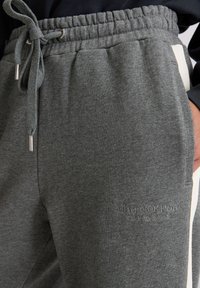 Close-up van grijze sweatpants met elastische tailleband, touwtje, zijzakken, een crèmekleurige streep en het geborduurde logo van "Marc O'Polo".