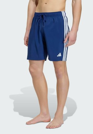Uomo che indossa pantaloncini sportivi blu con strisce bianche laterali e logo, in piedi a piedi nudi su uno sfondo semplice.