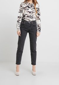 Schwarze Jeans mit hoher Taille und geradem Bein, kombiniert mit einer gemusterten Bluse in Creme-, Braun- und Schwarztönen. Weiße Schuhe mit Absatz.