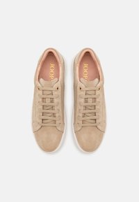 Beige semskinnsko med rosa innersåler, med flate lisser, gulldetaljer, og en hvit gummisåle. Rektangulær form med en glatt tekstur.