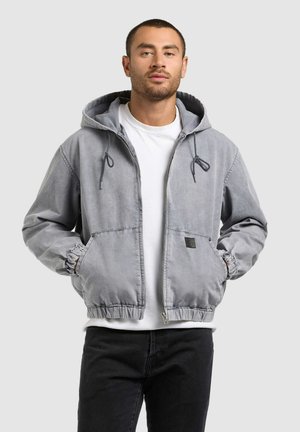 Vyras dėvi šviesiai pilką užtraukiamą džemperį su kišenėmis, baltą marškinėlį ir juodas kelnes, stovi priešais paprastą šviesią foną.