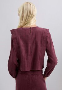 Blusa texturizada en color marrón con un diseño corto, que presenta hombros con volantes y un cierre de botones en la parte posterior. Tiene mangas largas.