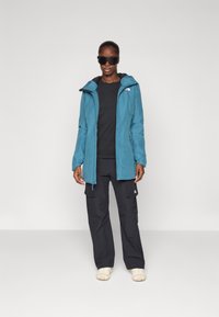 Teal vanntett jakke med justerbar hette, svart skjorte, cargo-bukser og lette sko. Tilbehør inkluderer mørke solbriller. Minimalistisk design.