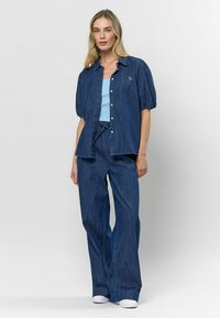 Džinsinė marškinė su trumpomis pūstomis rankovėmis, mėlynas tank topas ir platūs džinsiniai kelnių. Šviesiai mėlyni akcentai ir paprastas sagų priekyje detalė. Baltos sportbačiai.