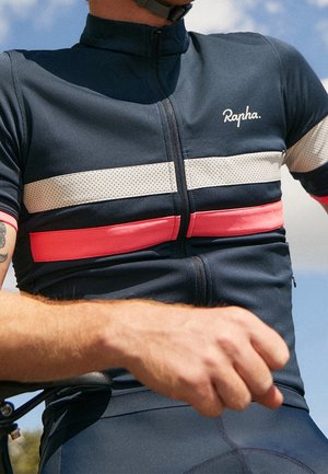 Kolarz w granatowej koszulce Rapha z białymi i różowymi paskami, widocznym tatuażem na ramieniu i zegarkiem na nadgarstku na tle częściowo zachmurzonego nieba.