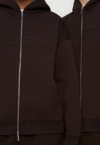 Deux sweat-shirts à capuche marron avec fermeture éclair, texte en relief sur la poitrine et tirettes de fermeture éclair argentées visibles.