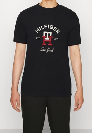 Osoba nosząca czarną koszulkę z logo "HILFIGER," "TH", "EST. 1985," i "New York" nadrukowanymi z przodu.