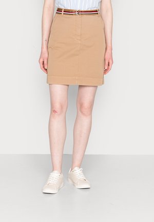 Osoba ubrana w beżową spódnicę do kolan z kieszeniami, paskiem w paski, białe trampki, stojąca na jednolitym jasnym tle.