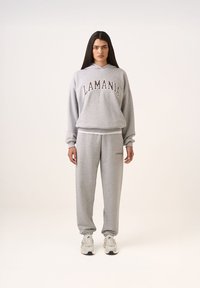 Szürke kapucnis pulóver és melegítőnadrág szett, amelyen sötétbarna színnel van feliratozva a "LAMANIA". Az ujjak és a bokák bordásak, a fazon pedig laza. Cipője fehér sportcipő.