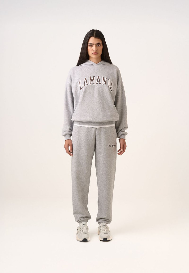 Szürke kapucnis pulóver és melegítőnadrág szett, amelyen sötétbarna színnel van feliratozva a "LAMANIA". Az ujjak és a bokák bordásak, a fazon pedig laza. Cipője fehér sportcipő.