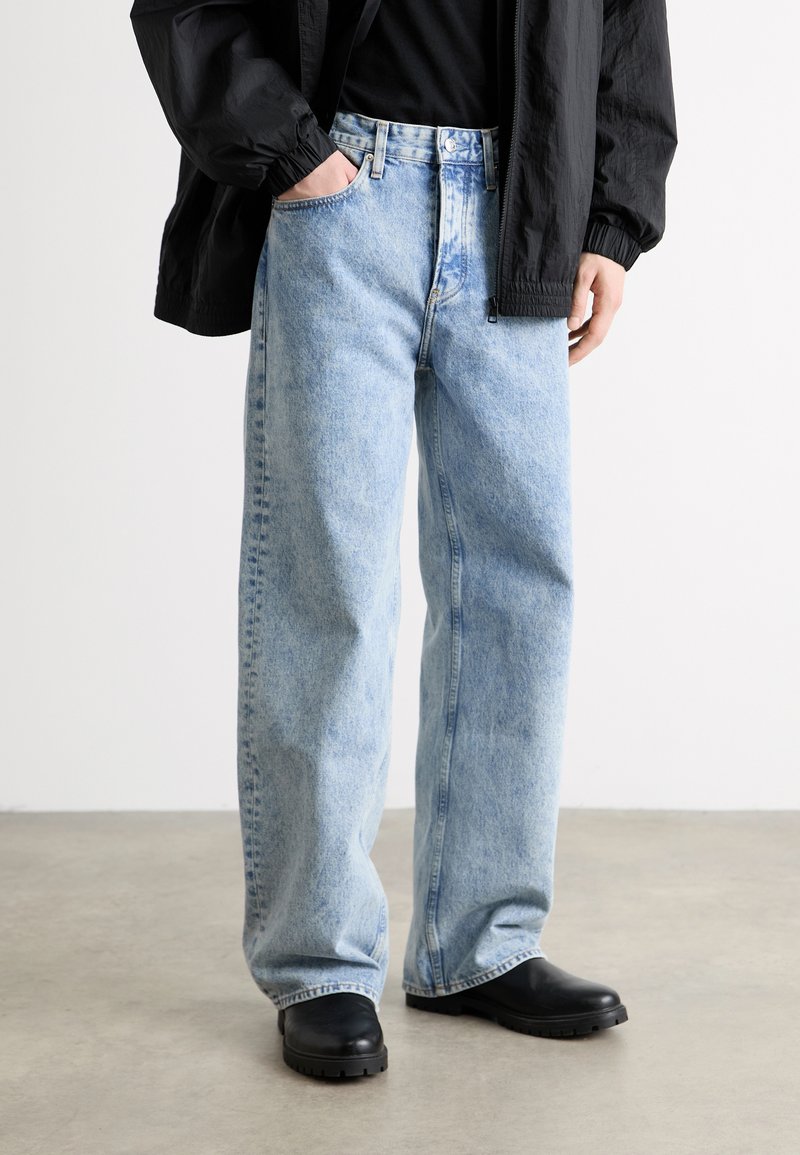 Light blue denim jeans with a relaxed fit, featuring five pockets and a zip fly. Paired with black rubber-soled ankle boots.