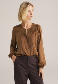 Brun, långärmad blus med plisserad halsringning och knäppning framtill. Tyget har en slät yta. Bärs med mörkfärgade, skräddarsydda byxor.