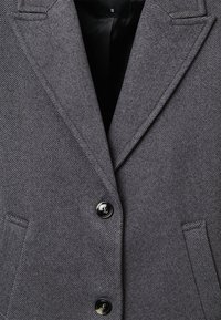 Blazer grigio a spina di pesce con revers, due bottoni neri e due tasche frontali a filetto, taglia piccola visibile sull'etichetta interna nera.