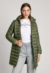 Olivgrön puffjacka med horisontell quiltad mönster, huva, framdragkedja och sidofickor, bärs över en vit grafisk t-shirt och ljusa jeans.