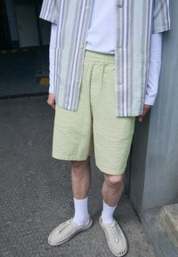Pantalones cortos de textura verde claro combinados con una camisa de botones de manga corta a rayas en tonos pastel. Unos zapatos tejidos de color crema y calcetines blancos completan el conjunto.