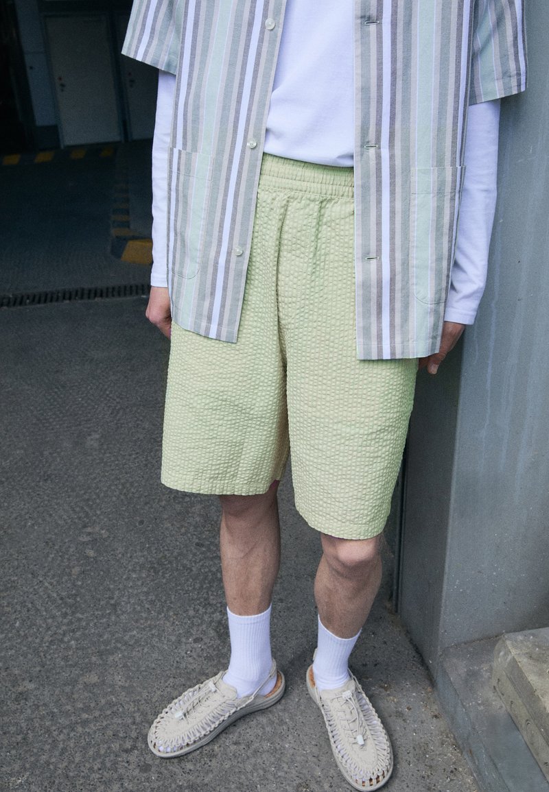 Pantalones cortos de textura verde claro combinados con una camisa de botones de manga corta a rayas en tonos pastel. Unos zapatos tejidos de color crema y calcetines blancos completan el conjunto.