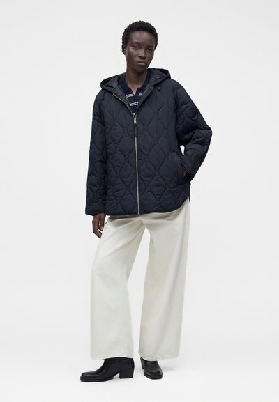Personne portant une veste matelassée noire à capuche, une chemise rayée, un pantalon blanc à jambes larges et des bottines noires, debout contre un fond uni.