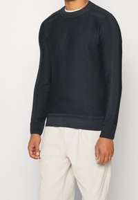 Donkerblauwe trui met een gestructureerde breiwijze, ronde halslijn en een subtiel logo op de borst. Heeft ribgebreide mouwen en zoom, gecombineerd met lichte broeken.