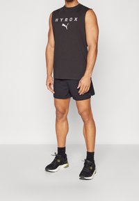 Tenue de sport pour homme comprenant un débardeur noir avec le texte blanc "HYROX" et un short noir. Complétée par des baskets noires et des chaussettes foncées.
