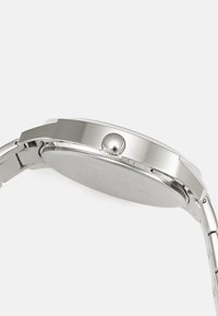 Zegarek ze stali nierdzewnej z okrągłą kopertą, polerowanym wykończeniem oraz koroną po boku. Metalowe ogniwa bransolety i gładki design tylnej części.