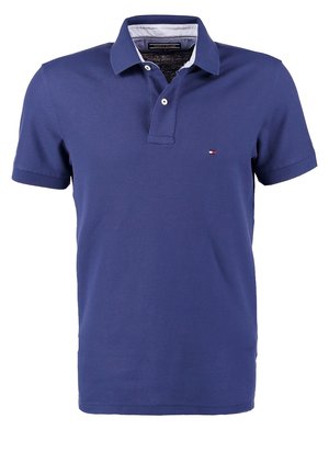 Personne debout, les mains dans les poches, portant une veste beige, un polo bleu, un pantalon noir et des baskets blanches sur un fond uni.
