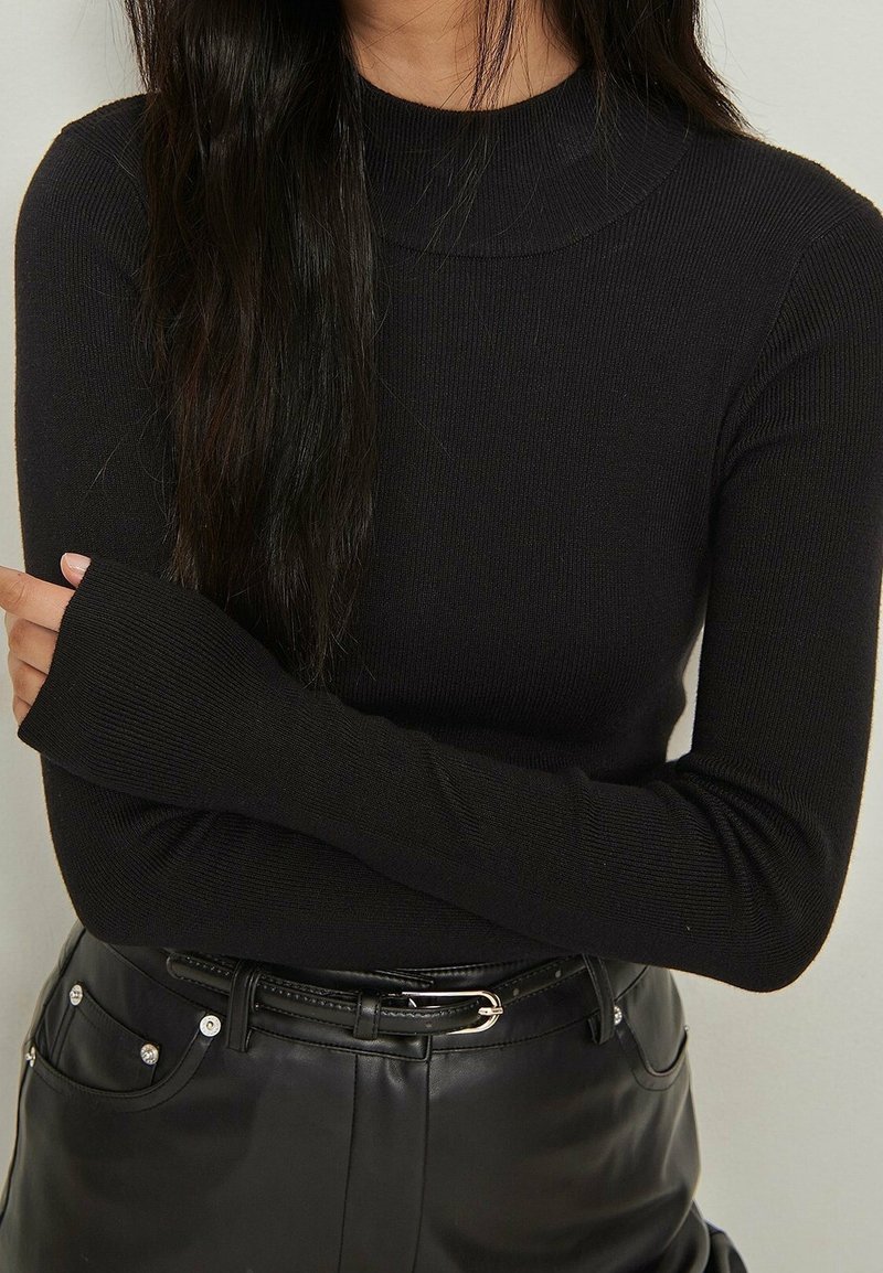Femme portant un haut noir côtelé à manches longues rentré dans un pantalon en cuir noir taille haute avec une fine ceinture noire et une boucle argentée.