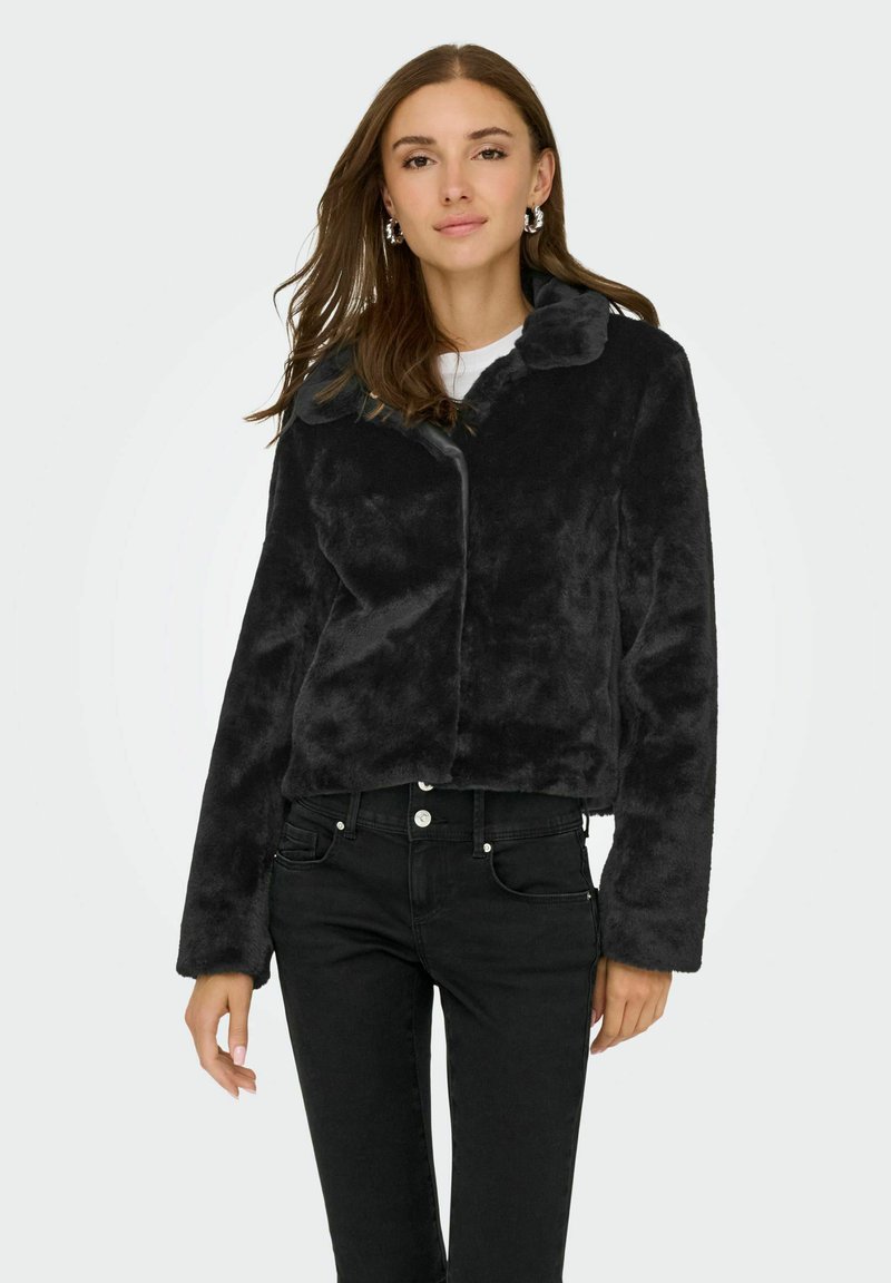 Femme portant une veste en fausse fourrure noire avec col, des boucles d'oreilles argentées en forme de cercle et un jean noir taille haute, se tenant devant un fond clair uni.