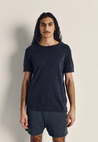 Ung man med långt mörkt hår och mustasch står mot en enkel ljus bakgrund, klädd i en svart T-shirt och mörka shorts.