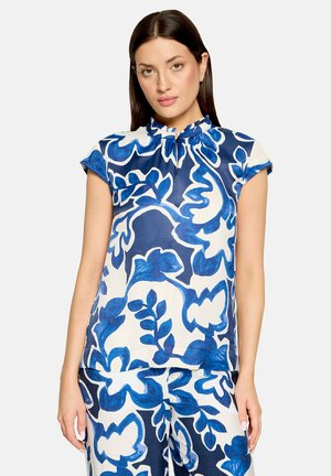 Femme portant un chemisier assorti à motifs floraux bleus et blancs avec manches courtes et col à volants, ainsi qu'un pantalon avec le même motif.