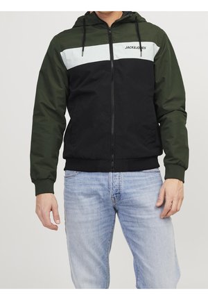 Mand iført grøn, sort og hvid Jack & Jones lynlåshættejakke med lyseblå jeans foran ensfarvet baggrund.