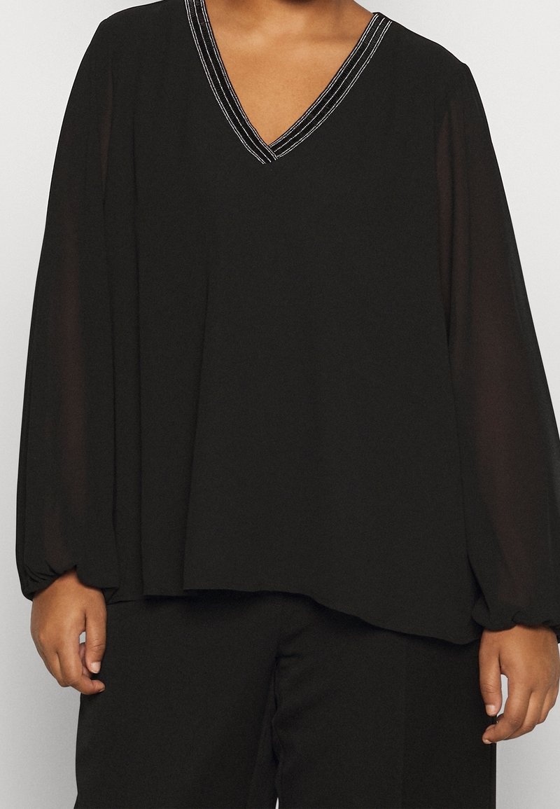Personne portant un chemisier noir ample avec de longues manches transparentes et un décolleté en V avec des détails de couture blancs, associé à un pantalon noir.