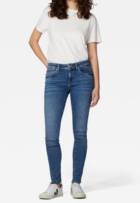 Jeans ajustados de color azul, material de mezclilla, cinco bolsillos, con un ligero desvanecimiento. Combinados con una camiseta blanca lisa y zapatillas claras.