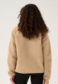 Beige fleecová bunda s texturovaným povrchem, zaoblenými rameny a dlouhými rukávy, bez viditelných zapínání nebo vzorů.